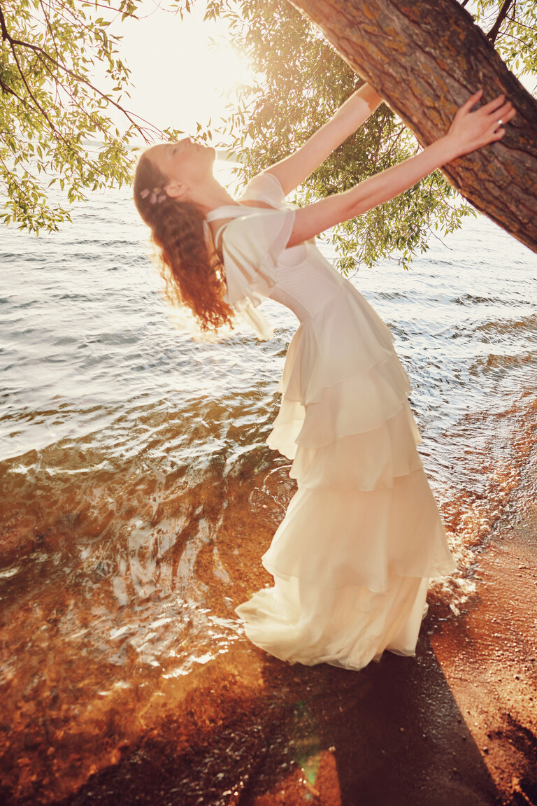 Side view of Sia off-white dress with voluminous tiered chiffon skirt posing near water