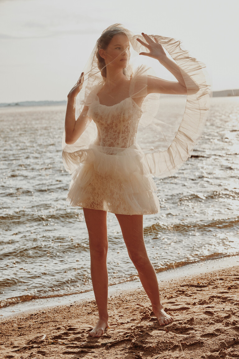 Bride holding veil wearing Luna short lace dress on the beach barefoot