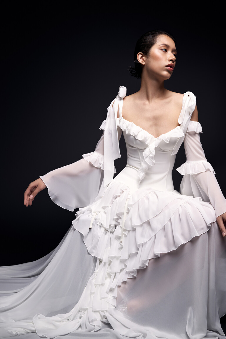 Seated model in Riot off-white gown showing voluminous ruffled skirt and sheer sleeves