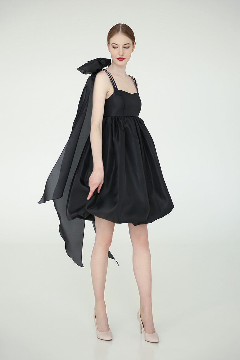Full body view of Leoni short black homecoming dress in organza with a statement shoulder bow and babydoll silhouette.