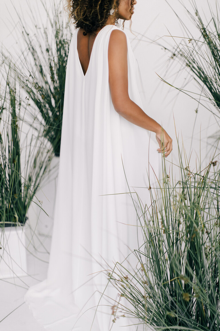 Model in the Unona Joan ivory dress, back view. The sleeveless gown features a deep V-cut back and a flowing cape-like effect.