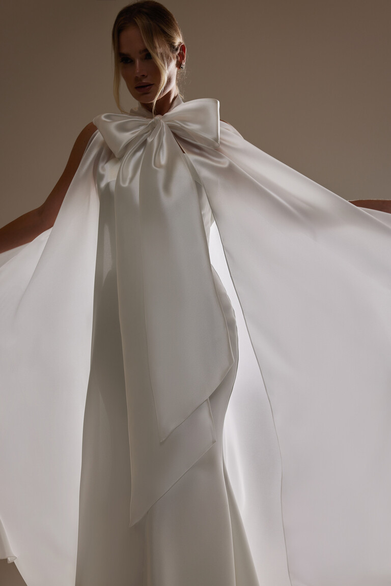 Unona Cape Casey ivory bridal cape over a fitted dress, highlighting the sheer organza panels and dramatic oversized bow.