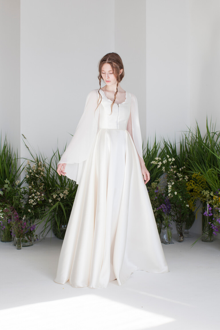 Styled full length view of Edite romantic ivory satin wedding dress with chiffon cape sleeves in a natural floral setting