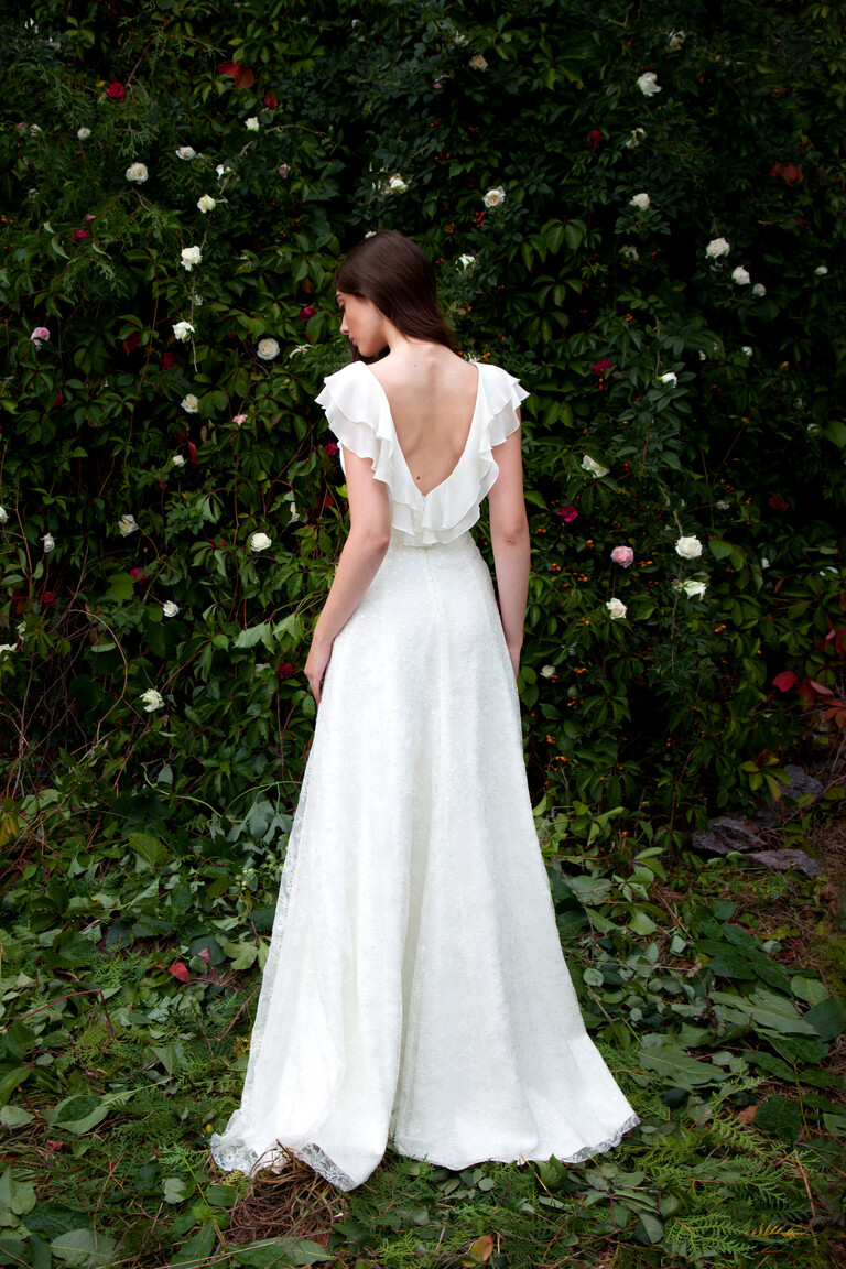 Back view of the Alison boho bridal gown in a garden setting, showcasing a breathtaking low V-back and floral lace skirt.