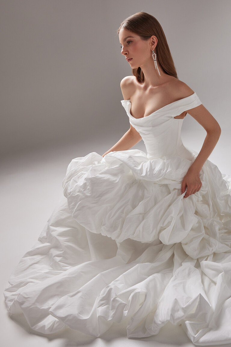 Paradise Off-Shoulder Taffeta Wedding Dress Ruffled Skirt Crouching Pose Unona Crouching pose in Paradise off-white taffeta bridal gown by Unona. Showcasing the off-shoulder bodice and ruffled skirt.