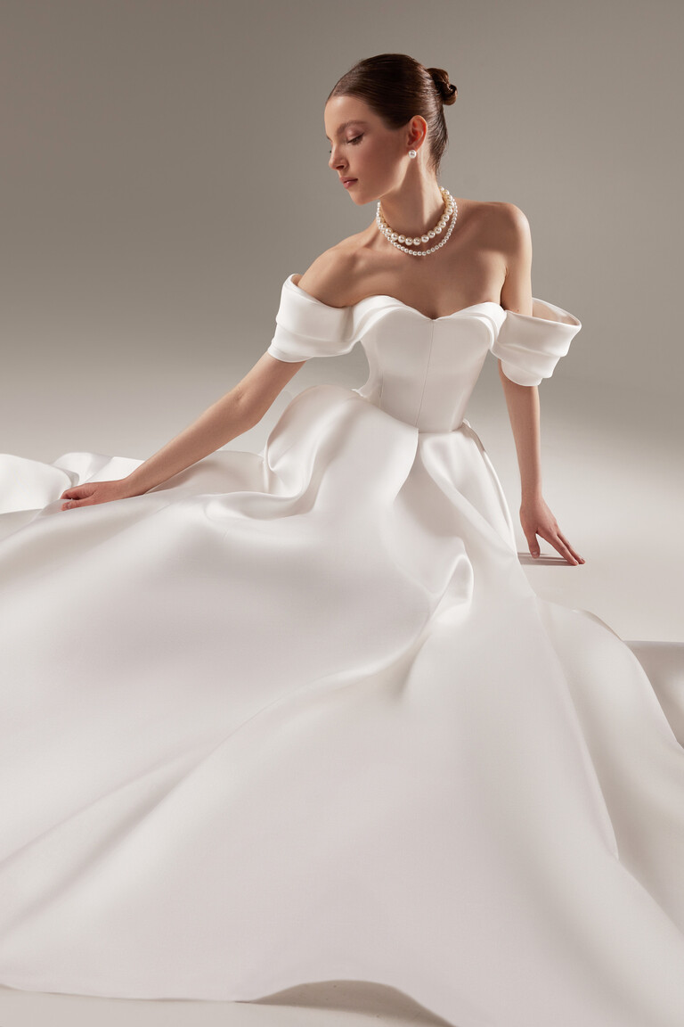 Romantic Seated View of Fly Ball Gown Seated bride wearing the romantic Fly off-the-shoulder ball gown in off-white Mikado with an architectural sweetheart bodice.